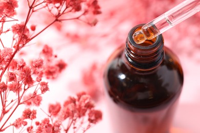 Photo of Dripping serum from pipette into bottle and gypsophila flowers on pink background, closeup