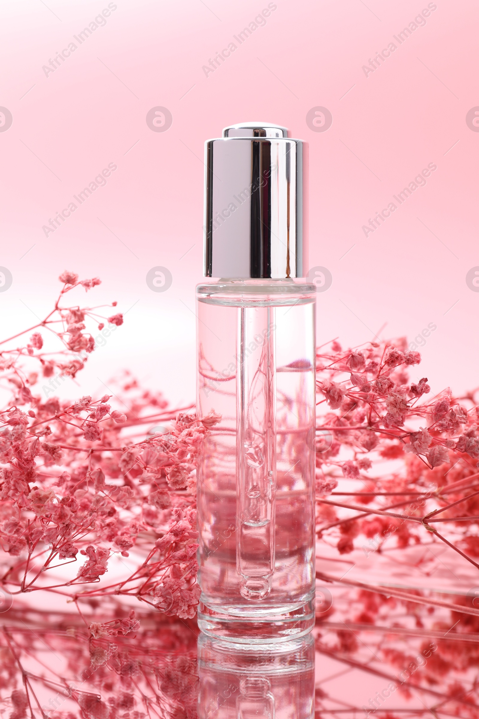 Photo of Bottle of serum and gypsophila flowers on mirror surface against pink background