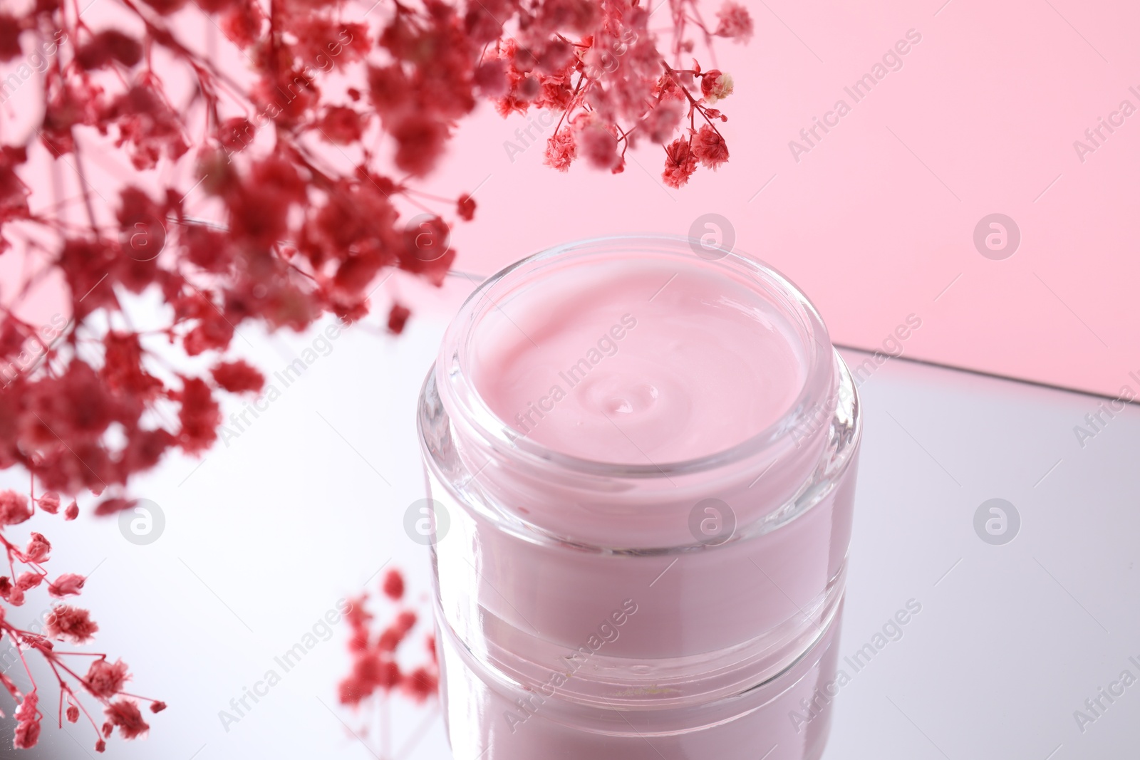 Photo of Jar of cream and gypsophila flowers on mirror surface against pink background, closeup