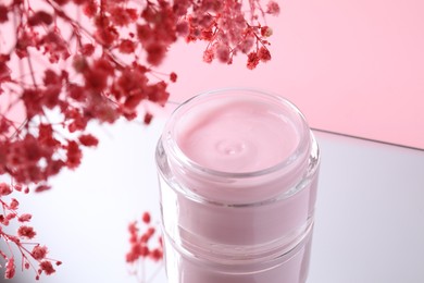 Photo of Jar of cream and gypsophila flowers on mirror surface against pink background, closeup