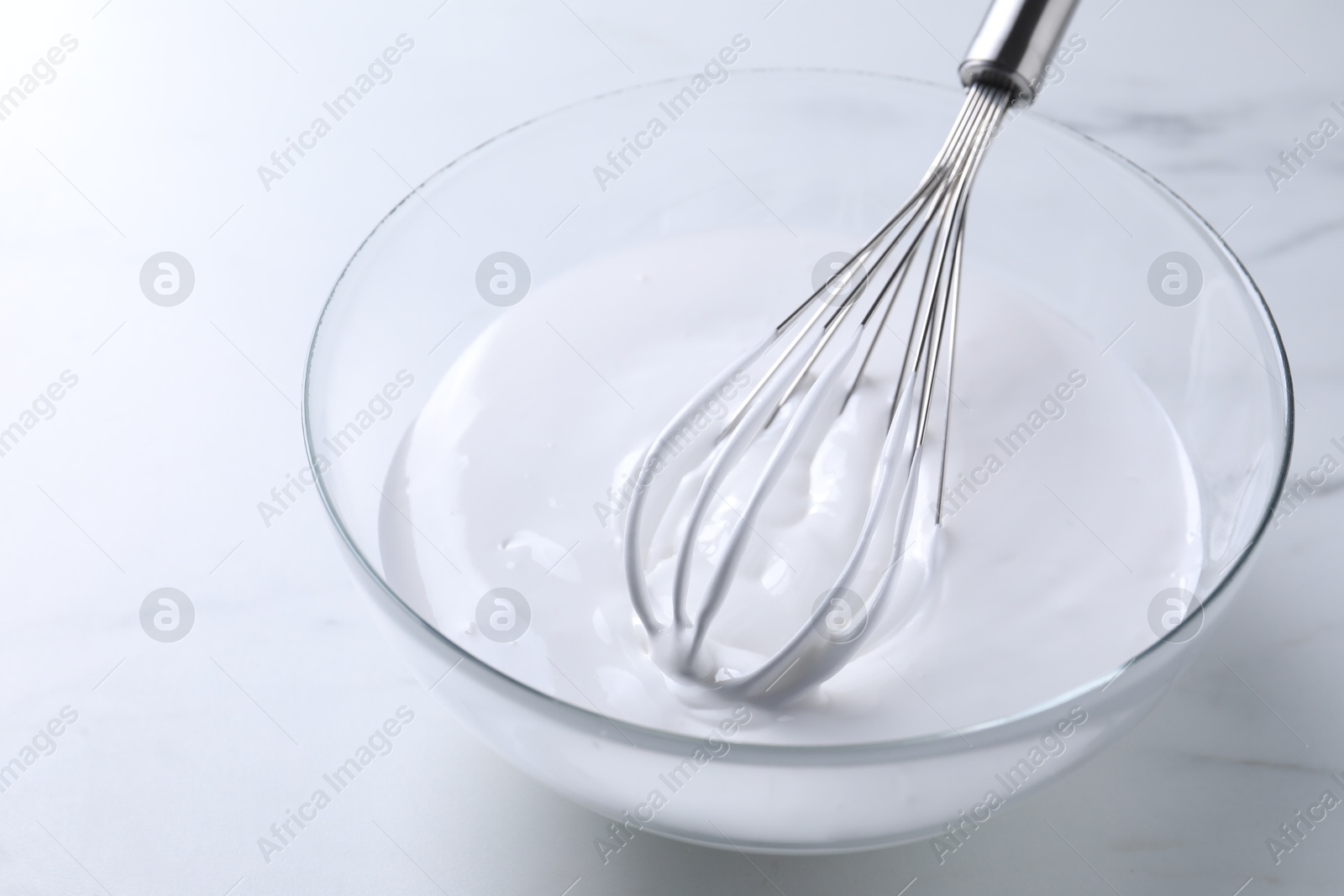 Photo of Whisk and whipped cream in bowl on white marble table, closeup