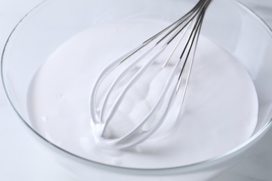 Photo of Whisk and whipped cream in bowl on white table, closeup