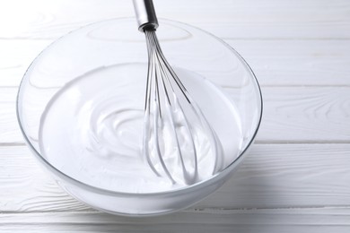 Photo of Whisk and whipped cream in bowl on white wooden table, closeup