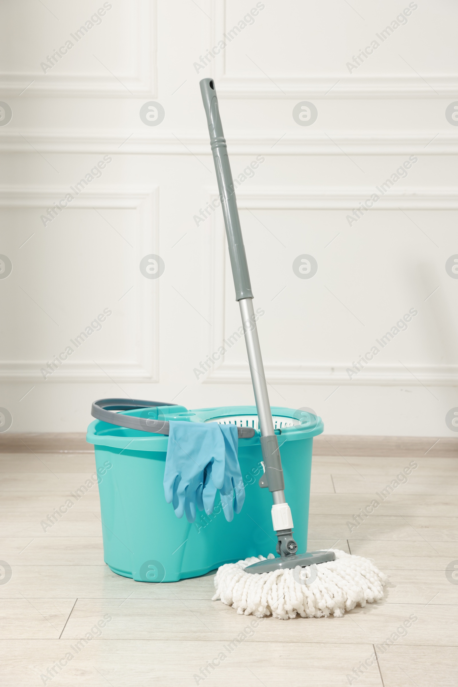 Photo of String mop, bucket and gloves on wooden floor near white wall