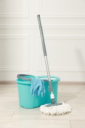 Photo of String mop, bucket and gloves on wooden floor near white wall