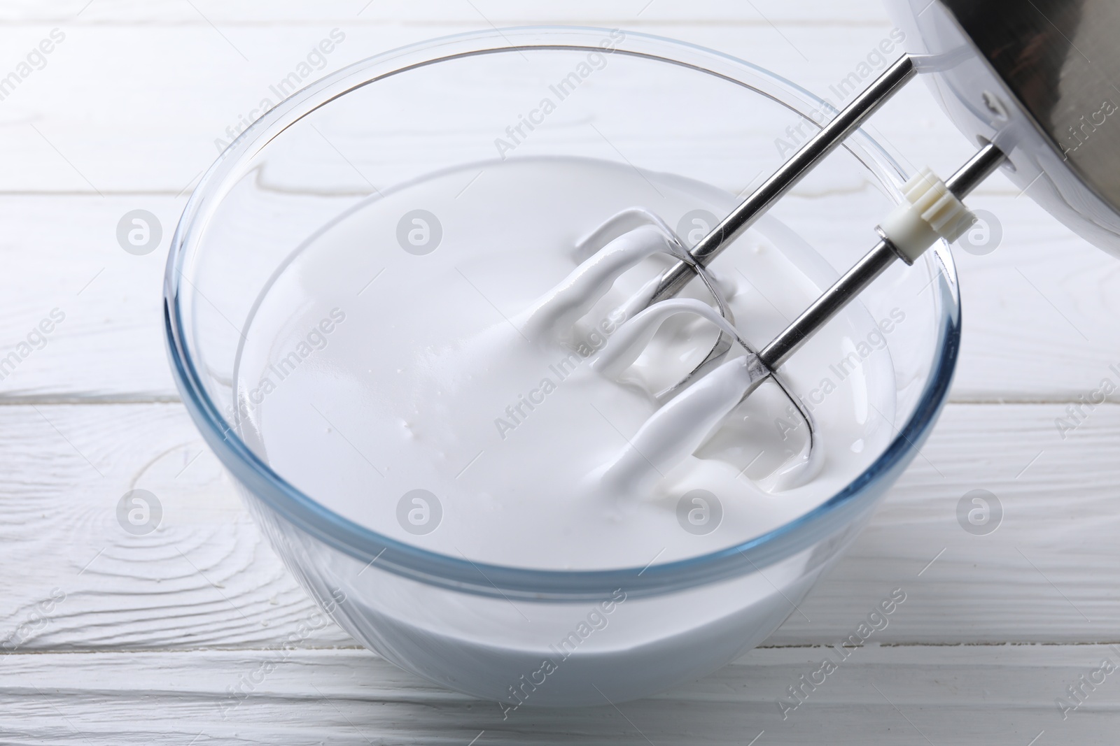 Photo of Mixing whipped cream with hand mixer in bowl on white wooden table, closeup