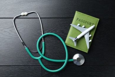 Photo of Medical tourism. Stethoscope, plane model and passport on black wooden table, flat lay