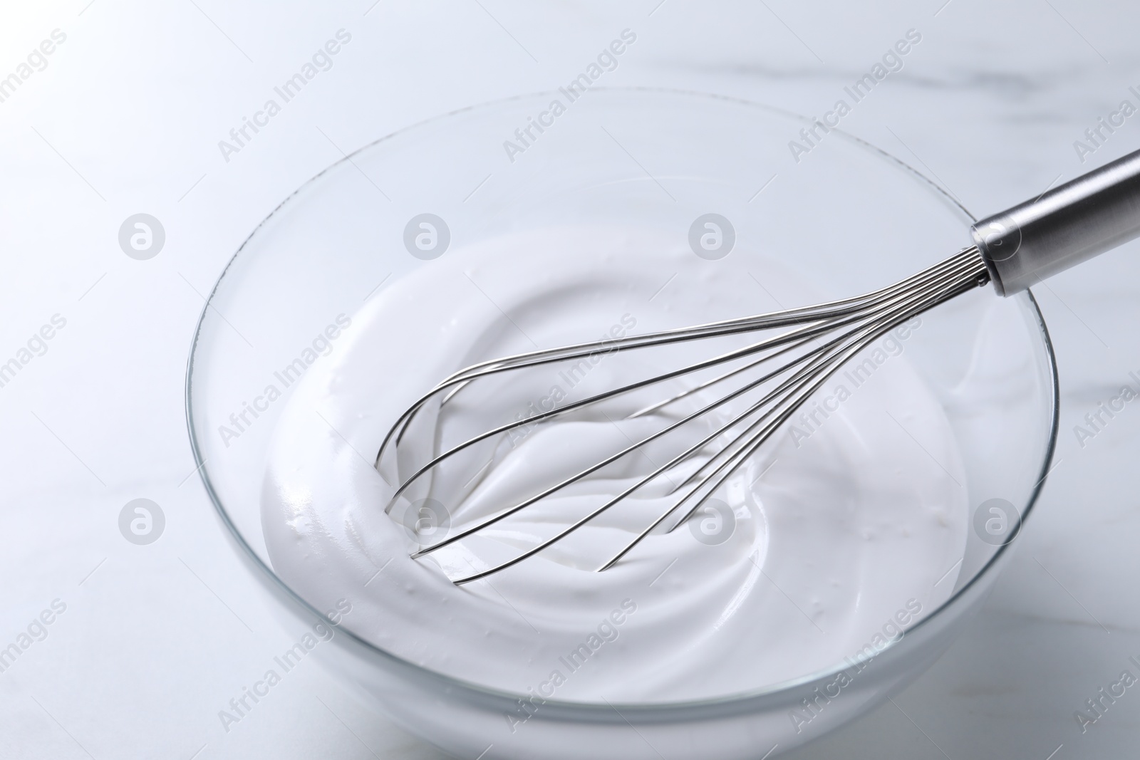 Photo of Whisk and whipped cream in bowl on white marble table, closeup