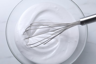 Photo of Whisk and whipped cream in bowl on white marble table, closeup