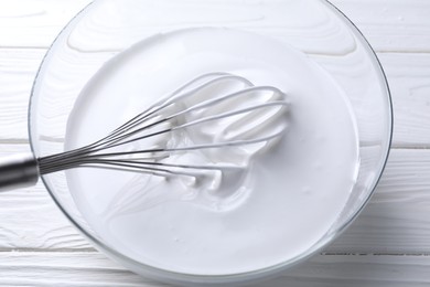 Photo of Whisk and whipped cream in bowl on white wooden table, closeup