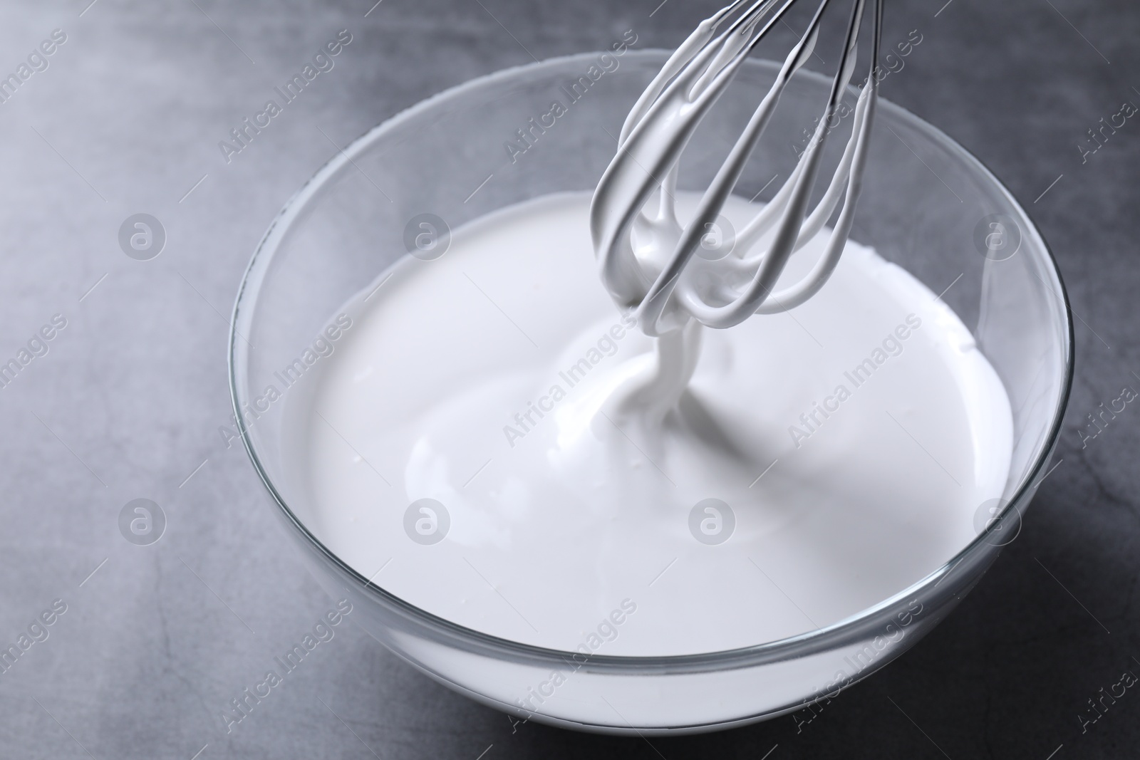 Photo of Mixing whipped cream in bowl with whisk on grey table, closeup