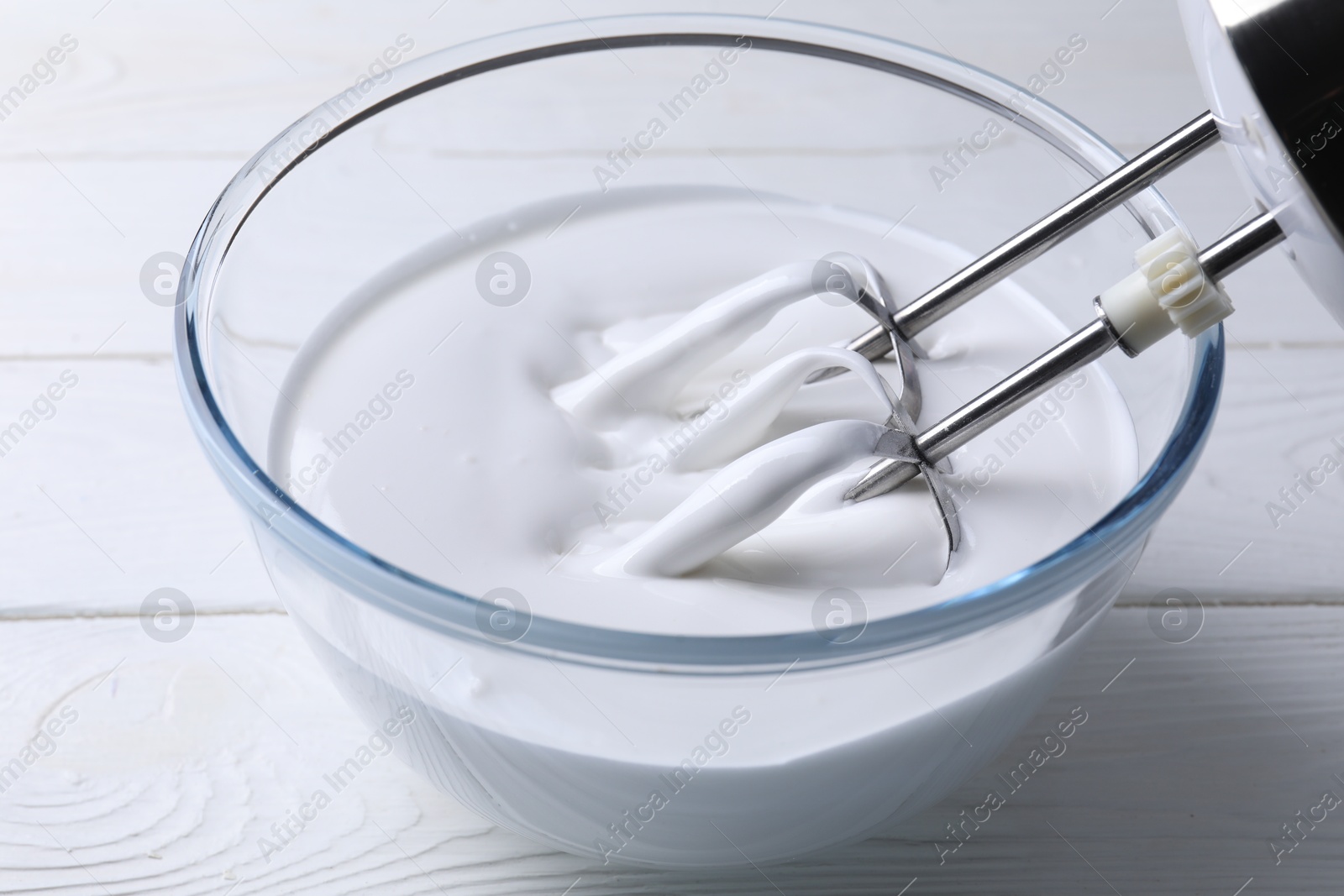 Photo of Mixing whipped cream with hand mixer in bowl on white wooden table, closeup