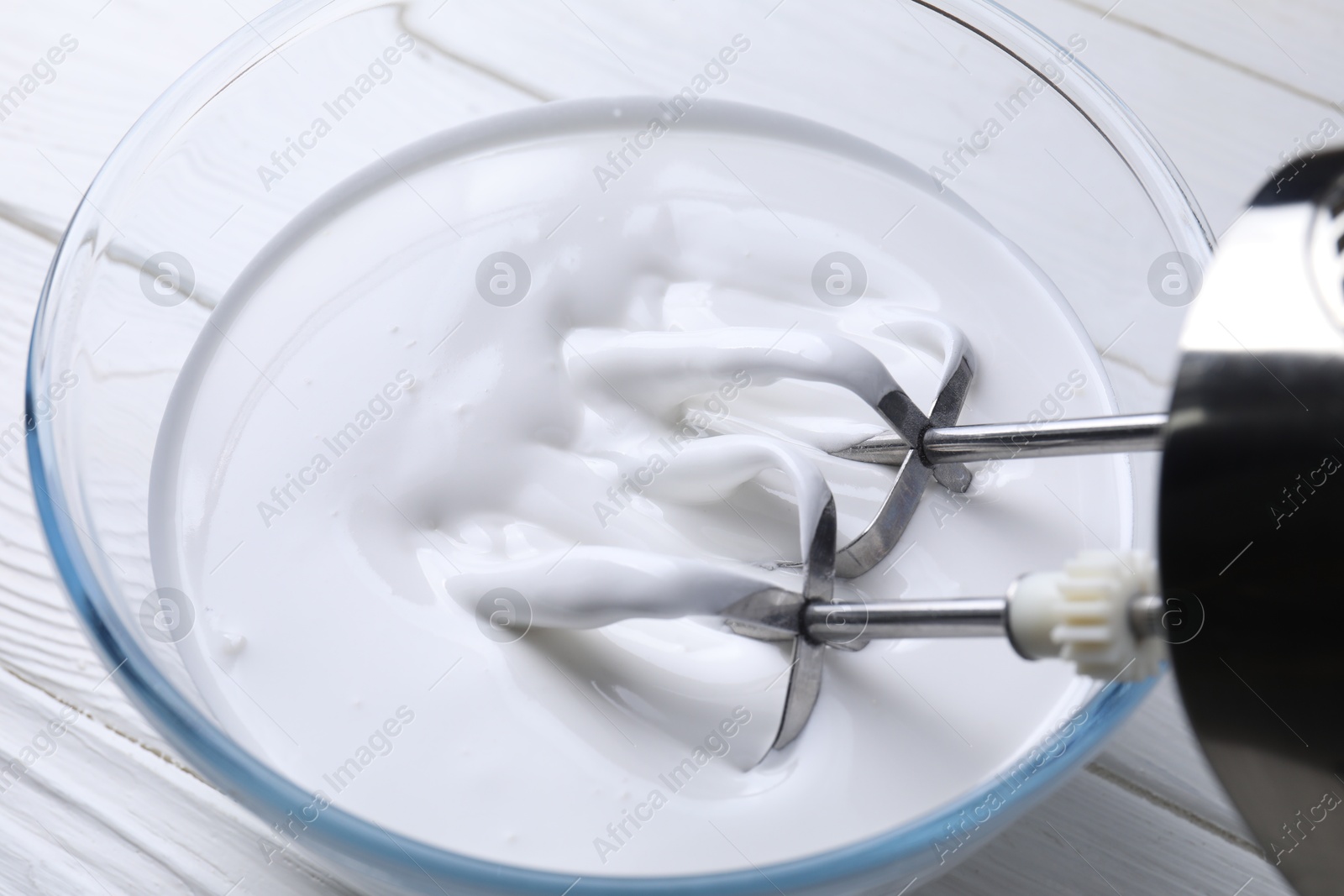 Photo of Mixing whipped cream with hand mixer in bowl on white wooden table, closeup