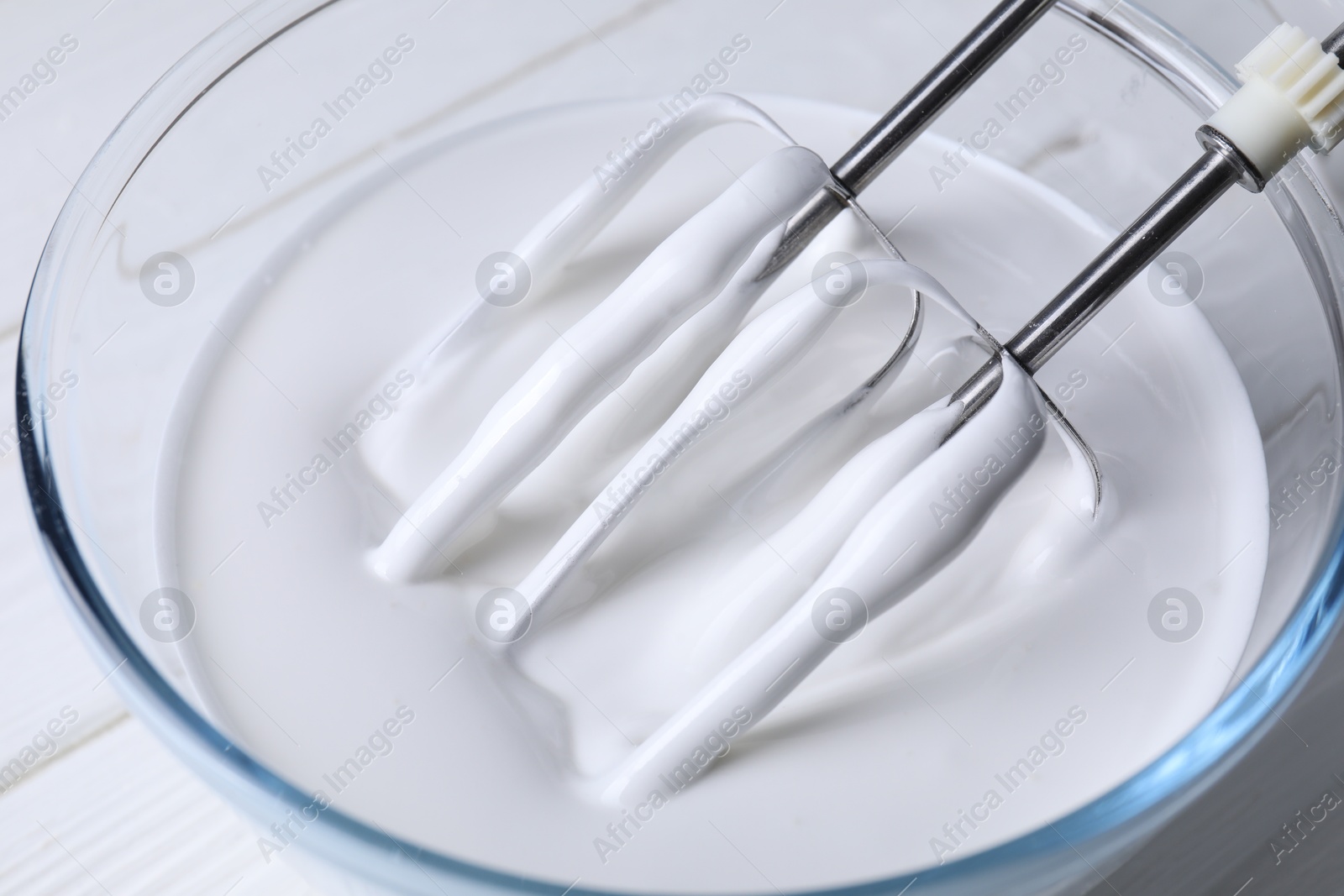 Photo of Mixing whipped cream with hand mixer in bowl on white wooden table, closeup