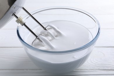 Photo of Mixing whipped cream with hand mixer in bowl on white wooden table, closeup