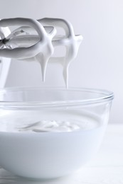 Photo of Hand mixer with whisks and bowl of whipped cream on white wooden table, closeup