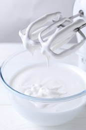 Photo of Hand mixer with whisks and bowl of whipped cream on white wooden table, closeup