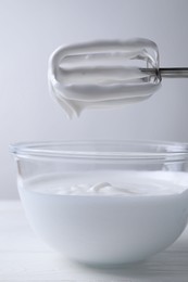 Photo of Hand mixer with whisks and bowl of whipped cream on white wooden table, closeup