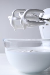 Photo of Hand mixer with whisks and bowl of whipped cream on white wooden table, closeup