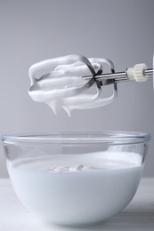 Photo of Hand mixer with whisks and bowl of whipped cream on white wooden table, closeup