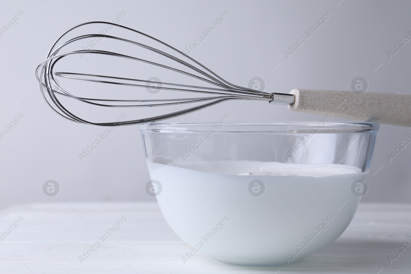 Photo of Whisk and bowl of whipped cream on white wooden table, closeup