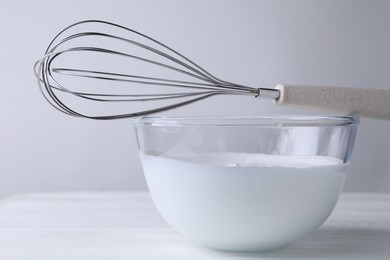 Photo of Whisk and bowl of whipped cream on white wooden table, closeup