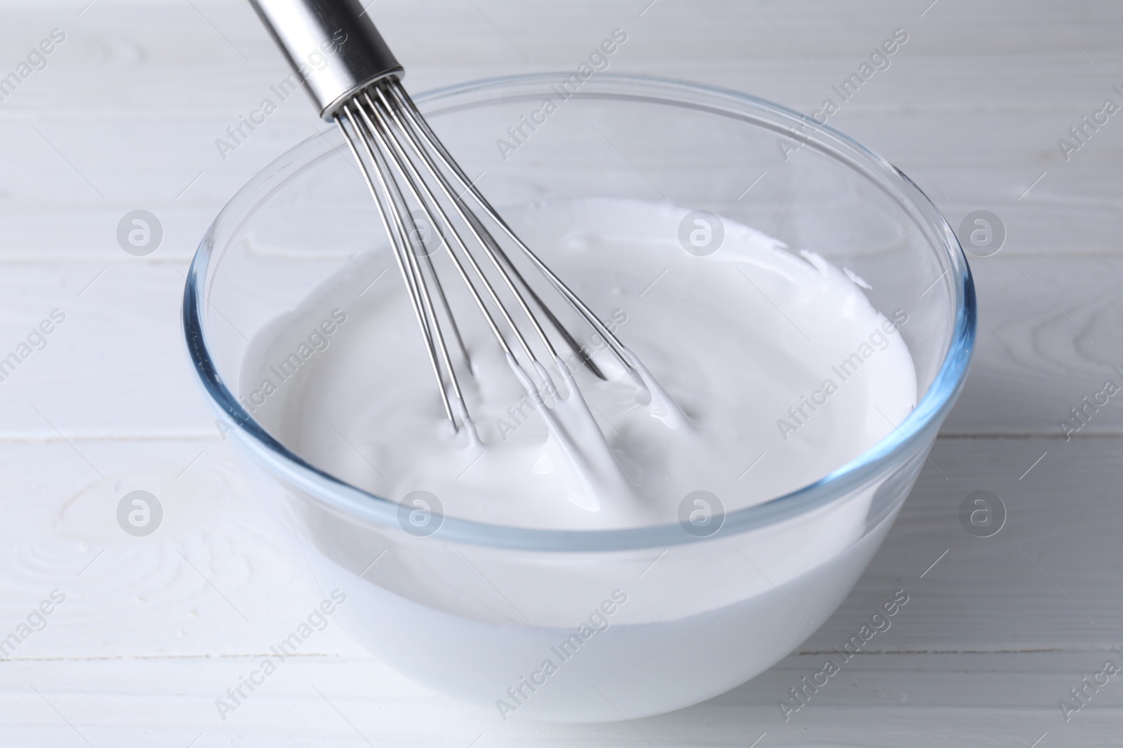 Photo of Whisk and bowl of whipped cream on white wooden table, closeup