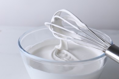 Photo of Whisk and bowl with whipped cream on white table, closeup