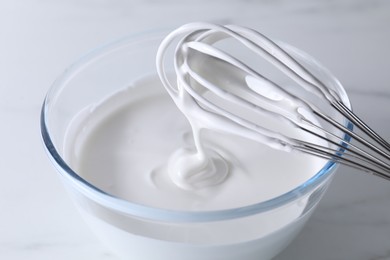 Photo of Whisk and bowl with whipped cream on white table, closeup