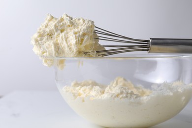 Photo of Whisk and bowl with whipped cream on white table, closeup