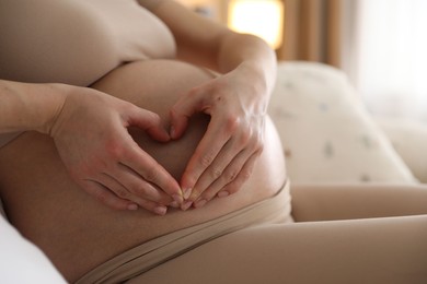 Pregnant woman making heart with hands on her belly at home, closeup