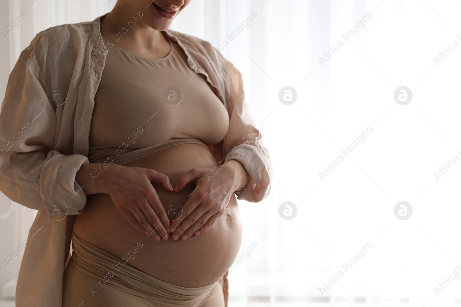 Photo of Pregnant woman making heart with hands on her belly near window at home, closeup. Space for text