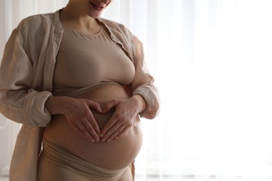 Pregnant woman making heart with hands on her belly near window at home, closeup. Space for text