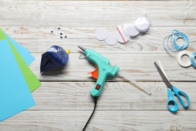 Photo of Hot glue gun and handicraft materials on white wooden table, flat lay