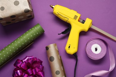 Photo of Hot glue gun and handicraft materials on purple background, flat lay