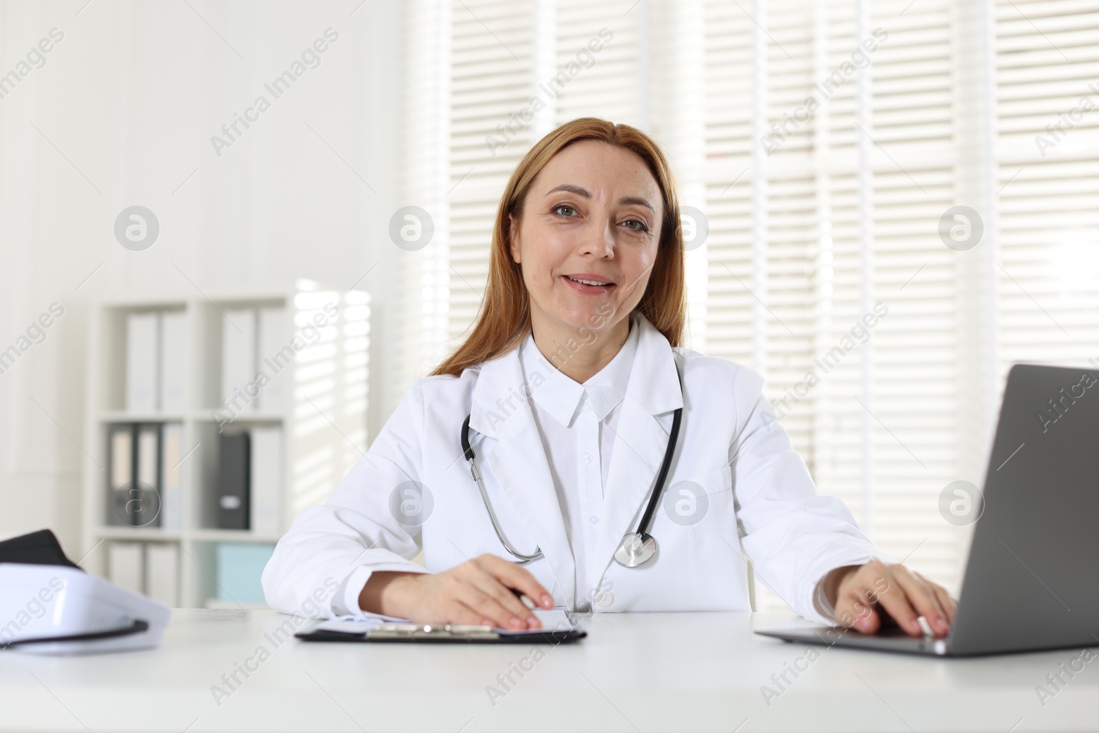 Photo of Cardiologist with stethoscope using laptop at white table in clinic