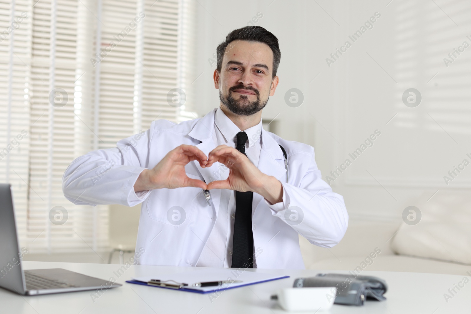 Photo of Cardiologist making heart with hands at white table in clinic