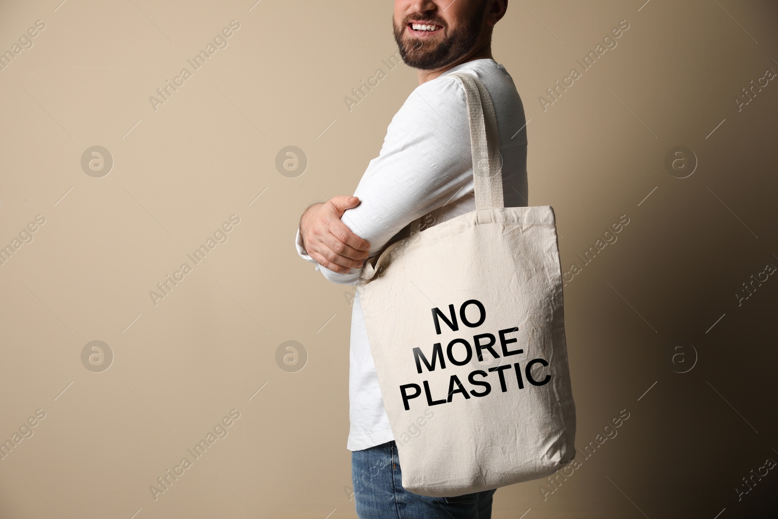 Image of Happy young man holding eco bag with No More Plastic text on dark beige background, closeup