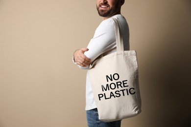 Happy young man holding eco bag with No More Plastic text on dark beige background, closeup