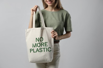 Image of Happy young woman holding eco bag with No More Plastic text on light grey background, closeup