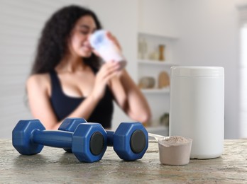 Protein powder and dumbbells on table in room where woman drinking protein shake, selective focus