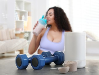 Protein powder and dumbbells on table in room where woman drinking protein shake, selective focus