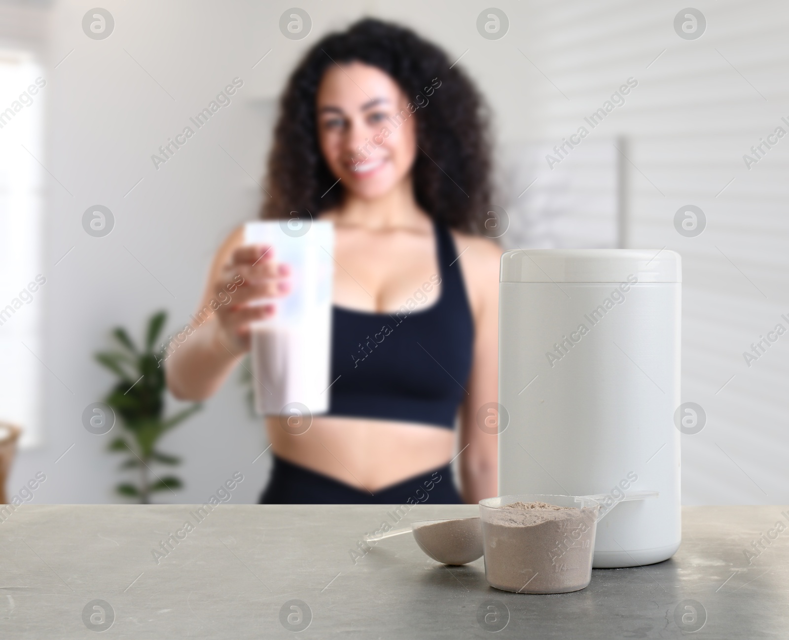 Image of Protein powder on table in room where woman holding protein shake, selective focus