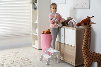 Photo of Little girl standing on step stool and playing with toys indoors