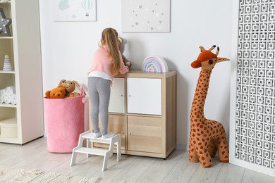 Photo of Little girl standing on step stool and playing with toys indoors