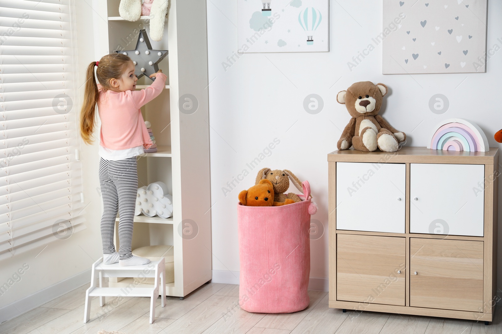 Photo of Little girl standing on step stool and reaching for toys on shelf indoors