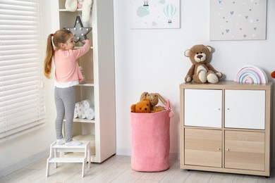 Photo of Little girl standing on step stool and reaching for toys on shelf indoors