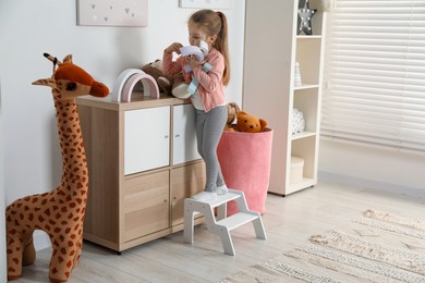 Photo of Little girl standing on step stool and playing with toys indoors