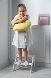 Photo of Little girl with toy standing on step stool indoors