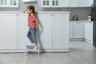 Photo of Little girl standing on step stool and reaching towards counter in kitchen, space for text